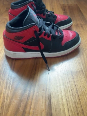 Air Jordan 1 Mid svart/röd - Säljer ett par Air Jordan 1 Mid i klassisk svart och röd färg med vita sulor. Skorna har svarta skosnören, perforerad tåbox och ikoniska Jordan-loggan på sidan. Tillverkade i läder och textil för en snygg och bekväm look.