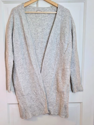 Ljusgrå lång kofta - By Malene Birger - Merino & kashmir - Mjuk och lyxig kofta från By Malene Birger i modell Altina. Ljusgrå, öppen design i merinoull och kashmirblandning. Perfekt som varm lager-på-lager-kofta året runt.  - Storlek Medium - Den är längre i modellen (ca 85 cm från axeln), med rymlig passform över bysten (ca 51 cm ärmhål–ärmhål).  - Praktiska fickor på sidorna och den typiska BM Birger-lyxkänslan i strukturen. - Nypris var cirka 3300 kr.  Skick: Nästan som ny och inget märkbart slitage, visst ytludd finns som är oundvikligt med kashmir