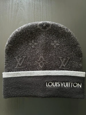 Mörkgrå/svart Louis Vuitton mössa - Snygg mörkgrå mössa från Louis Vuitton med diskret monogrammönster och ljusgrå kant. Märkets logga är broderad framtill. Perfekt accessoar för dig som vill ha en exklusiv touch till din outfit. Tillverkad i mjukt stickat material.