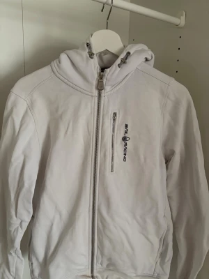 Vit hoodie från Sail Racing - Säljer en vit hoodie från Sail Racing med dragkedja framtill och en bröstficka med logga. Hoodien har justerbar huva och är tillverkad i mjukt material, perfekt för en avslappnad och sportig stil.