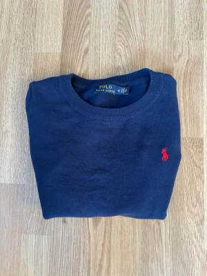 Mörkblå stickad tröja Polo Ralph Lauren - Mörkblå stickad tröja från Polo Ralph Lauren med rund halsringning och den klassiska röda logotypen broderad på bröstet. Tröjan har långa ärmar och ribbade muddar vid ärmslut och nederkant. Perfekt för en stilren och avslappnad look. Pris kan diskuteras.