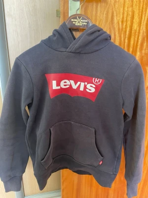 Svart hoodie från Levi's med logga - Säljer en blå  hoodie från Levi's med den klassiska röda loggan framtill. Hoodien har en stor känguruficka och huva. Tillverkad i mjukt bomullsmaterial och har ribbade muddar vid ärmar och nederkant. Perfekt för en avslappnad och cool stil.Storlek 12a (152cm). Passar alltså personer som är runt 150cm. Fint skick inga fläckar eller skador. 