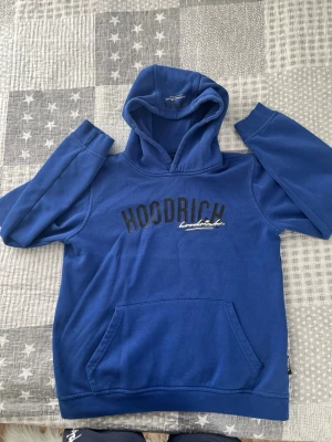 Blå hoodie från Hoodrich - Snygg blå hoodie från Hoodrich med svart och vit logga på bröstet och tryck på ryggen. Klassisk känguruficka framtill och stor huva. Tillverkad i mjukt bomullsmaterial, perfekt för en avslappnad streetwear-look. Storlek 12-13y/ L