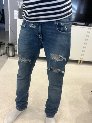 Blå slitna jeans från Polo Ralph Lauren - Säljer ett par blå jeans från Polo Ralph Lauren med slitningar och hål på både fram- och baksida. Jeansen har raka ben, klassisk femficksmodell och coola detaljer på bakfickorna. Perfekta för en avslappnad och lite unikate look . Riktigt bra skick men de har slitningar för utseendets skull 