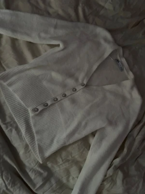 Vit kofta med pärlknappar från Zara - Mysig vit kofta från Zara med v-ringning och dekorativa pärlknappar framtill. Koftan har ribbade muddar vid ärmslut och nederkant samt lång ärm. Perfekt för lager på lager och enkel att styla till olika outfits.