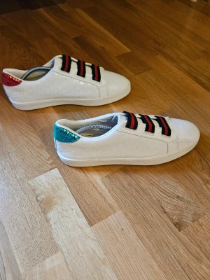 Gucc Ace vita sneakers med färgglada detaljer - Säljer ett par vita Gucci Ace sneakers i läder med klassiska gröna och röda ränder på elastiska band istället för snören. Baksidan har metallicdetaljer i rött på ena skon och grönt på den andra. Diskret GG-mönster i lädret ger extra lyxig känsla.