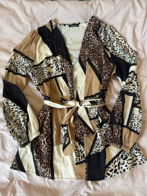 Mönstrad blus från Boohoo med knytband - Snygg omlottblus från Boohoo med leopardmönster i beige, svart och brunt. Blusen har v-ringning, långa ärmar med resår vid handlederna och ett matchande knytband i midjan. Tillverkad i ett lätt och mjukt material som ger en skön känsla.