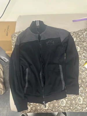 Svart ziptröja från BOSS - Snygg svart ziptröja från BOSS med grå detaljer på axlar och fickor. Tröjan har hög krage, dragkedja framtill och två fickor. Perfekt för en stilren och avslappnad look.