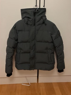 Mörkgrå pufferjacka med huva - Snygg mörkgrå pufferjacka med stor huva och dragkedja framtill. Jackan har två sidofickor, ribbade muddar vid ärmsluten och en patch på ena ärmen. Perfekt för kalla vinterdagar och riktigt skön att bära. Materialet är slitstarkt och håller värmen bra. Använd cirka 4 gånger 