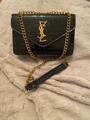 Svart YSL handväska med kedja - Säljer en svart handväska från Saint Laurent med croco-mönstrat läder och guldfärgad kedja. Väskan har det klassiska YSL-emblemet framtill och en axelrem i kedja med läderdetalj. Perfekt för dig som gillar lyxiga accessoarer och stilren design.