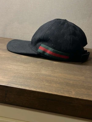 Svart Gucci keps med GG-mönster - Snygg svart keps från Gucci med klassiskt GG-monogram i tyget och en grön-röd rand på sidan. Kepsen har böjd skärm och justerbar rem bak. Tillverkad i canvasmaterial som ger en exklusiv känsla och streetig look.