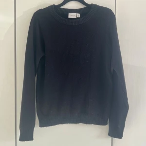Svart stickad tröja från VILA - En enkel och stilren svart stickad tröja från VILA. Tröjan har rund halsringning, långa ärmar och ribbade muddar vid ärmslut och nederkant. Perfekt att matcha med jeans eller kjol för en clean look.