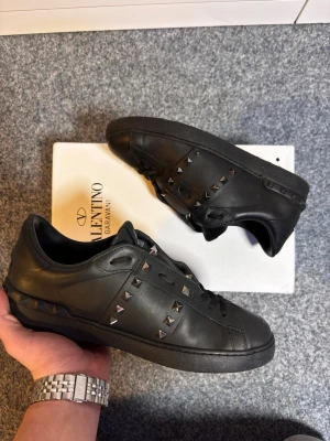 Svarta Valentino skor  - Säljer ett par exklusiva Valentino Rockstud Untitled (modell nr 11) i helsvart läder. Skorna är i ett mycket fint begagnat skick och är väl omhändertagna. En tidlös sneaker som kombinerar lyx med en modern stil. • Modell: Rockstud Untitled / No. 11. • Storlek: 41 (Serienummer/storlek bekräftat på sista bilden). • Färg: Svart (Nero) med matchande svarta nitar (ruthenium/gunmetal look). • Skick: Bra skick. Mindre bruksspår finns på sulan och tån, men inget som stör helhetsintrycket. • Äkthet: 100%