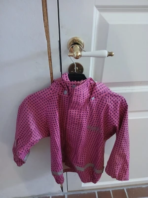 Rosa regnjacka med mönster och byxor - Säljer en rosa vindjacka med mörkblå fyrkantigt mönster och reflexdetaljer. Jackan har huva, dragkedja framtill och resår vid ärmslut och nederkant. Perfekt för blåsiga dagar och enkel att ta på. Materialet är troligtvis polyester. (Står namn)
