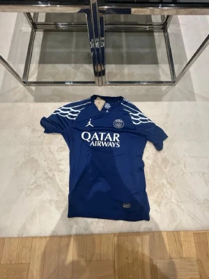 Blå PSG matchtröja Jordan x Qatar - Snygg blå PSG matchtröja i samarbete med Jordan och Qatar Airways. Tröjan har vita detaljer på axlarna, klubbmärke och sponsortryck framtill samt namn och nummer (D. Doué 14) på ryggen. Tillverkad i lätt och funktionellt material, perfekt för fotboll eller träning.