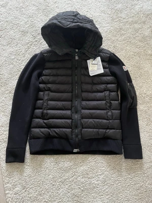 Svart cardigan från Moncler - Snygg svart cardigan från Moncler med quiltad front och huva. Ärmarna är stickade för extra komfort och jackan har dragkedja framtill samt två fickor. Moncler-logga på ärmen och diskreta detaljer gör den riktigt stilren. Jag har aldrig använt den. All plast och taggar sitter kvar. Jag tar byten. Kom dm för mer info eller bilder.