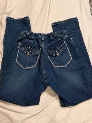 Mörkblå jeans med strassdetaljer - Snygga mörkblå jeans med raka ben och resår i midjan. Baksidan har två fickor dekorerade med strass och silverfärgade nitar samt kontrastsömmar. Perfekta för dig som vill ha lite extra bling i vardagen.