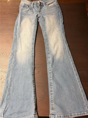 Ljusblå  lågmidjande bootcut jeans - Säljer ett par ljusblå bootcut jeans med snyggt slitna detaljer och broderade bakfickor. Jeansen har klassisk femficksmodell, normal midja och vida ben. Perfekta för dig som gillar retrostil och vill sticka ut lite extra.