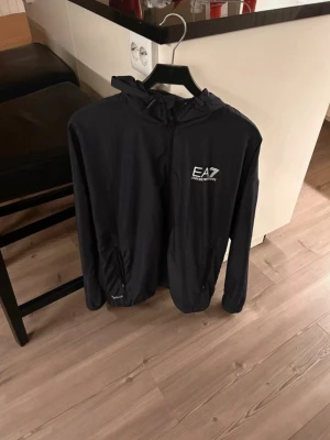 Svart vindjacka från Emporio Armani - Snygg svart vindjacka från Emporio Armani EA7 med huva och dragkedja framtill. Jackan har två fickor med dragkedja och en vit logga på bröstet. Perfekt för dig som vill ha en stilren och sportig look. Den har en fläck på framsidan, som går att ta bort OBS