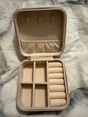 Beige smyckesväska i sammet - Snygg och kompakt smyckesväska i beige sammet med flera fack och ringrullar. Perfekt för att organisera ringar, örhängen och halsband. Fyrkantig form och mjuk insida som skyddar dina accessoarer. Enkel att ta med i väskan eller på resan.