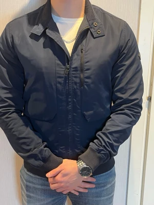 Mörkblå J.Lindberg vindjacka med krage - Stilren mörkblå vindjacka med klassisk krage och dragkedja framtill. Jackan har två framfickor, ribbade muddar vid ärmslut och midja samt en diskret patch på ärmen. Materialet är slätt och lätt, perfekt för blåsiga dagar.