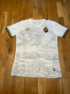 Vit AIK special kit från Nike - Säljer en vit AIK matchtröja från Nike med svart och guld detalj på ärmsluten. Framsidan har ett snyggt tryck av Stockholmssilhuett och AIK:s klubbmärke på bröstet. Tillverkad i lätt och ventilerande material, perfekt för träning eller match.