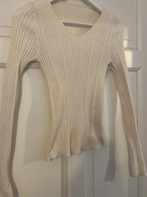 Vit ribbstickad tröja med v-ringning - Säljer en vit ribbstickad tröja med v-ringad hals och långa ärmar. Tröjan har figurnära passform och bred ribbad kant nedtill. Perfekt för dig som gillar en enkel men snygg stil. Materialet känns mjukt och stretchigt.
