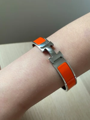 Orange H-klikarmband från H - Snyggt armband från H i silverfärgad metall med orange emaljdetaljer. Armbandet har den ikoniska H-formade låset och en stilren, modern design som verkligen sticker ut. Perfekt accessoar för dig som vill addera färg och lyx till din outfit.