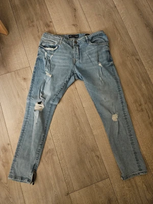 Ljusblå slitna jeans - Säljer ett par ljusblå skinny fit jeans med slitningar på både lår och knän. Jeansen har klassisk femficksmodell och dragkedjor vid bensluten för extra edge. Endast använd 2-3 gånger pga fel storlek
