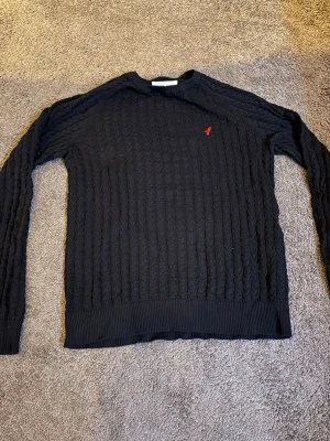 Marinblå kabelstickad tröja med röd logga - Säljer en Marinblå kabelstickad tröja med rund hals och ribbade muddar. Tröjan har en liten röd broderad logga på bröstet och är långärmad. Perfekt för dig som gillar klassisk stil med en twist.