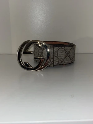 bälte med silverfärgat spänne - Snyggt bälte i beige och grått med klassiskt mönster och stort, silverfärgat spänne. Bältet är gjort i skinn och har exklusiv känsla, perfekt för att lyfta vilken outfit som helst.