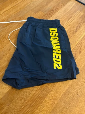 Marinblå DSQUARED2 badbyxor - Snygga marinblå badbyxor från DSQUARED2 med stort gult logotryck längs ena benet. Klassisk kort modell med resår i midjan och vit dragsko för bästa passform. Tillverkade i lätt och snabbtorkande material, perfekta för stranden eller poolen.