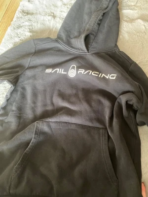 Svart hoodie från Sail Racing - Svart hoodie från Sail Racing med stor vit logga på bröstet. Klassisk modell med huva och magficka. Tillverkad i mjukt bomullsmaterial som känns skönt mot huden. Perfekt för dig som gillar en sportig och avslappnad stil.