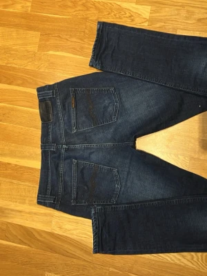 Mörkblå jeans från Nudie Jeans  - Säljer ett par mörkblå jeans från Nudie Jeans med klassisk femficksmodell och raka ben. Jeansen har snygga slitningar och kontrastsömmar, samt en patch bak i midjan. Perfekta för dig som gillar stilrena och tidlösa jeans. Storlek W30 L32 Modell Lean Dean