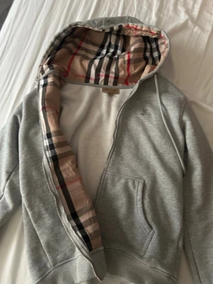 Grå hoodie från Burberry med dragkedja - Säljer en grå hoodie från Burberry med klassiskt rutigt foder i huvan och dragkedja framtill. Hoodien har fickor på sidorna och diskret Burberry-logga broderad på bröstet. Perfekt för dig som gillar stilrena och exklusiva plagg.