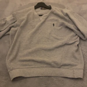 Grå v-ringad tröja från Polo Ralph Lauren - Klassisk grå v-ringad tröja från Polo Ralph Lauren med den ikoniska broderade loggan på bröstet. Tröjan har ribbade muddar vid ärmslut och nederkant, och är tillverkad i mjukt material som passar perfekt till lager på lager.