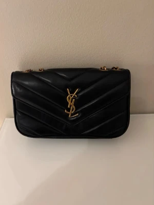 Svart YSL handväska i läder - Säljer en svart handväska från Yves Saint Laurent i mjukt läder med quiltat mönster. Väskan har guldfärgade detaljer och ikoniska YSL-loggan framtill. Kedjeremmar i guld ger en lyxig känsla och väskan har en klassisk, rektangulär form.