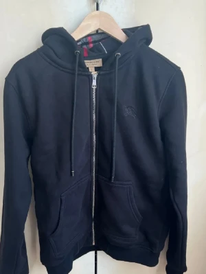 Svart zip hoodie från Burberry - Snygg svart hoodie från Burberry med dragkedja framtill, två stora fickor och klassisk huva med dragsko. Diskret Burberry-logga broderad på bröstet och typiskt rutigt foder i huvan. Perfekt för en chill och stilren look.