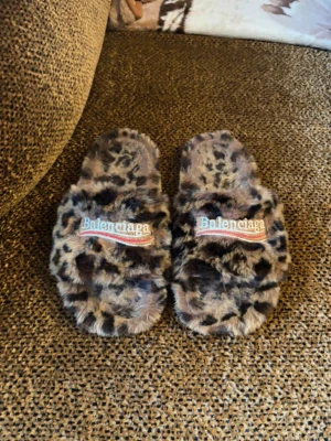 Balenciaga fur tofflor - Säljer ett par fluffiga tofflor från Balenciaga med leopardmönster och broderad logga på ovansidan. Tofflorna har öppen tå och mjuk, lurvig yta i syntetmaterial. Sulan är svart med Balenciaga-inpräglad text. Perfekta för dig som vill ha något extra hemma.