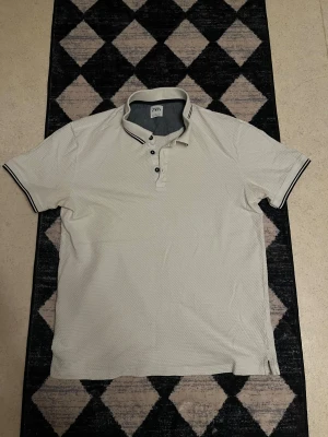Vit pikétröja från Zara - Säljer en vit pikétröja från Zara med klassisk krage och knappar framtill. Tröjan har diskreta svarta ränder på kragen och ärmarna samt en strukturerad yta. Perfekt till jeans eller shorts för en clean look.