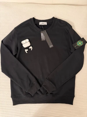 Stone Island sweater  - Snygg svart sweatshirt från Stone Island med klassisk rund hals och ribbade muddar. Tröjan har den ikoniska Stone Island-loggan på vänster ärm och är tillverkad i mjukt bomullsmaterial. Perfekt för en clean och stilren look.