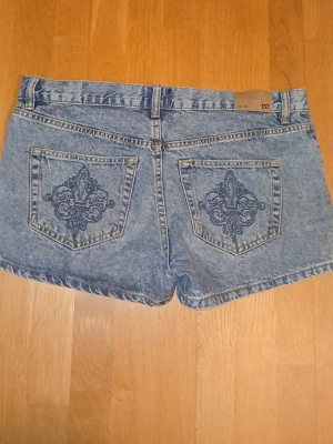 Blå jeansshorts - Säljer ett par blå högmidjade jeansshorts med broderad detalj på bakfickan. Klassisk femficksmodell i loose fit och snygg tvättad denim. Perfekta till sommaren och lätta att matcha med olika toppar.