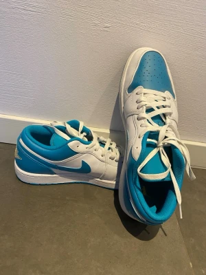 Nike Air Jordan 1 Low blå/vit - Säljer ett par Nike Air Jordan 1 Low sneakers i blått och vitt. Skorna har klassisk låg siluett, vit bas med blå detaljer och swoosh, samt guldbroderad Air Jordan-logga på hälen. Ovandelen är i läder och de har vita skosnören.