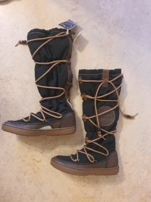 Svarta och bruna boots från Svea - Boots från Svea i svart textil med bruna detaljer och snörning runt hela skaftet. Grov sula i brunt gummi och förstärkta partier i läderimitation. Stora Svea-loggan på sidan och dekorativa träpärlor på snörena. Perfekta för kalla dagar.