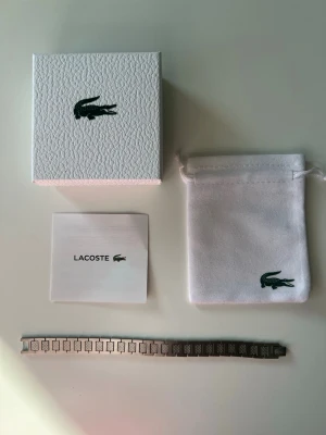 Silverfärgat armband från Lacoste - Snyggt armband i silverfärgad metall från Lacoste med klassisk krokodil-logga på spännet. Armbandet har rektangulära länkar med diskret mönster och kommer i originalförpackning med tygpåse och manual. Perfekt accessoar för en stilren look. Den är cirka 20cm lång men har en länkbortagare som kan göra den till din önskade storlek.