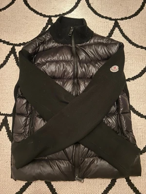 Svart dunväst från Moncler - Snygg svart dunväst från Moncler med stickade svarta ärmar och hög krage. Västen har dragkedja framtill med Monclers signaturband i rött, vitt och blått samt två sidofickor med dragkedja. Moncler-logga på ena ärmen. Perfekt för lager-på-lager och streetstyle.
