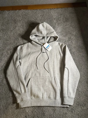 Grå hoodie från Nike med huva - Snygg grå hoodie från Nike med klassisk huva och dragsnören. Hoodien har en liten svart Nike-logga broderad på bröstet och en stor magficka framtill. Tillverkad i mjukt bomullsmaterial som känns skönt mot huden. Perfekt för en avslappnad och sportig stil.