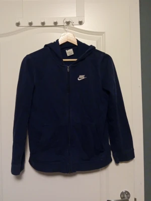 Mörkblå Nike hoodie med dragkedja - Snygg mörkblå hoodie från Nike med dragkedja framtill och vit Nike-logga på bröstet. Tröjan har huva och två fickor framtill. Perfekt för en avslappnad och sportig stil. Tillverkad i mjukt material för extra komfort.