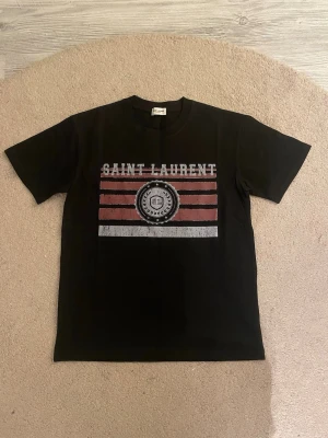 Saint Laurent t-shirt - Snygg svart t-shirt från Saint Laurent med tryck i vitt och rött framtill. T-shirten har korta ärmar, rund halsringning och raka linjer. Materialet känns mjukt och skönt, troligtvis bomull. Perfekt för dig som gillar streetstyle och designerplagg.