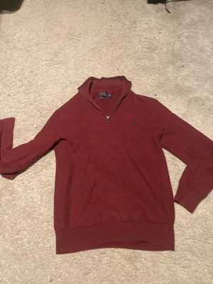 Röd halvzip tröja från Polo Ralph Lauren - Snygg röd tröja från Polo Ralph Lauren med halv dragkedja och broderad blå logga på bröstet. Tröjan har ribbade muddar och hög krage. Perfekt för lager på lager och en klassisk stil.